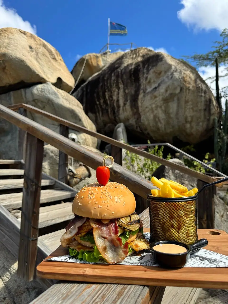 Burger on a wooden board with the Casibari rock formations and the Aruban flag behind